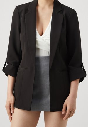 Person iført en sort blazer med opsmøgede ærmer over en hvid top med vrid foran og et kort gråt skørt.