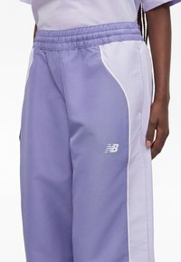 Άτομο που φοράει αθλητική παντελόνα New Balance σε λιλά και ανοιχτό μωβ χρώμα με ελαστική μέση και λευκό λογότυπο στο μηρό.
