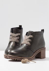 Bottines en cuir foncé avec talons épais et lacets en laine beige épais, placées sur une surface blanche sur un fond gris clair.