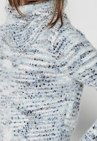 Pull en maille bleu clair avec un col drapé, présentant des nuances variées de bleu et de blanc, et une apparence texturée et duveteuse.