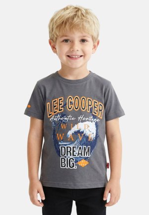 Jeune garçon blond souriant, portant un t-shirt gris Lee Cooper avec une grande vague bleue et le texte : Wild Wave Dream Big.