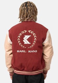 Bordowa kurtka varsity z kremowymi skórzanymi rękawami, z haftowanym logo i napisem "KARL KANI" na plecach, ze ściągaczami w paski na mankietach i dole kurtki.