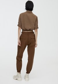 Femme vue de dos portant une chemise marron courte retroussée, un pantalon cargo marron taille haute et des bottines blanches à lacets sur un fond uni.
