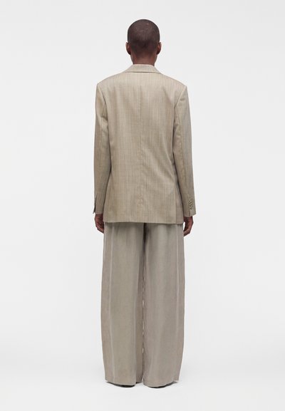 Personne debout, de dos, vêtue d'un blazer beige à carreaux et d'un pantalon beige à jambes larges, sur fond blanc uni.