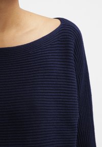 Námořnický modrý pletený svetr s širokým lodním výstřihem, který má výrazné žebrování a dlouhé rukávy, zdůrazňující uvolněný design.