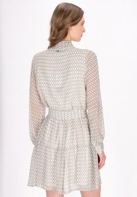 Langarm jurk van doorschijnende, lichte stof met een geometrisch patroon. Heeft een hoge hals, een cinched taille en een gelaagde rok.