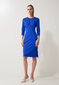 Robe bleue arrivant au genou avec des manches trois-quarts, présentant une accent en dentelle au niveau du décolleté et un détail en dentelle à l'ourlet. Texture de tissu lisse.