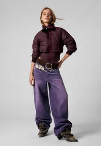 Korte gewatteerde jas in donkerpaars, met een hoge kraag en een ritssluiting aan de voorkant. Loszittende paarse spijkerbroek met een bloemenaccent en een riemdetail.
