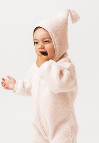 Malček, ki nosi mehak, svetlo roza flisov kombinezon s kapuco in cofkom, si v usta daje palec in gleda v stran na belem ozadju.