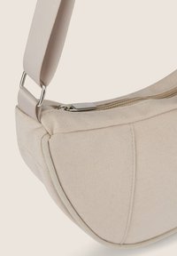 Borsa in tela beige con forma curvata, dotata di chiusura a zip e una robusta tracolla in tessuto fissata con accessori metallici.