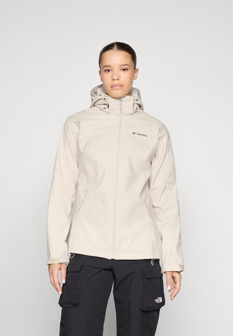 Veste softshell beige clair avec fermeture éclair, capuche et logo sur la poitrine ; comprend deux poches avant et des poignets ajustables.