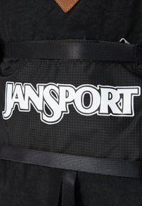 Sac à dos noir en tissu texturé, arborant un grand logo blanc "JANSPORT", un accent en cuir marron et des sangles en toile noires.