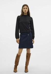 Haut noir à manches longues avec un col montant, associé à une jupe en denim foncé en forme de A, ornée de boutons dorés et de bottes brunes hauteur genoux.