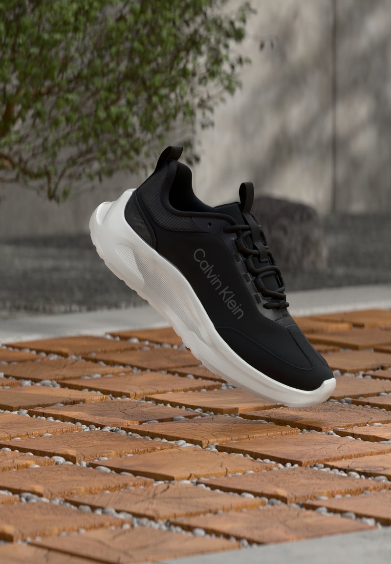 Chaussure de sport noire avec une texture lisse, semelle blanche et détail de logo ton sur ton. Comprend des lacets et une languette arrière. Flottant au-dessus de carreaux en bois.