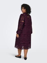 Spetsklänning i djup lila, med intrikata blomster mönster och en vågformad fåll. Långa ärmar med avslappnad passform.