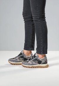 Schwarz-weiß gemusterte Sneaker mit strukturiertem Obermaterial, kontrastierenden schwarzen Akzenten und einer beigen Gummisohle, kombiniert mit eng anliegenden schwarzen Jeans.