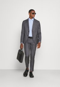 Completo grigio a quadri con vestibilità su misura, abbinato a una camicia azzurra e scarpe nere. Porta una cartella in pelle nera.
