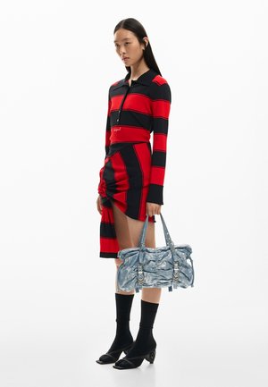 Žena oblečená v červeno-čiernom pruhovanom dlhom rukáve a sukni so záhybmi, držiaca textúrovanú svetlomodrú kabelku, s čiernymi ponožkami a lodičkami.