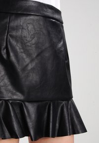 Gonna nera in ecopelle con vita aderente e orlo arricciato. Presenta una texture liscia e una leggera lucentezza. Lunghezza fino a metà coscia.