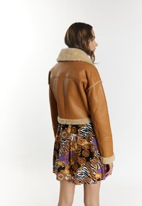 Brun læderjakke med en shearlingkrave og -manchetter, der har et cropped design. Kombinationen med en farverig mønstret nederdel.