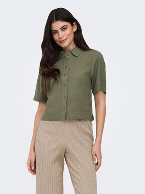 Pitkähiuksinen nainen, jolla on päällään napitettu oliivinvihreä lyhythihainen paita ja beiget, teksturoidut housut, seisomassa yksivärisen taustan edessä.