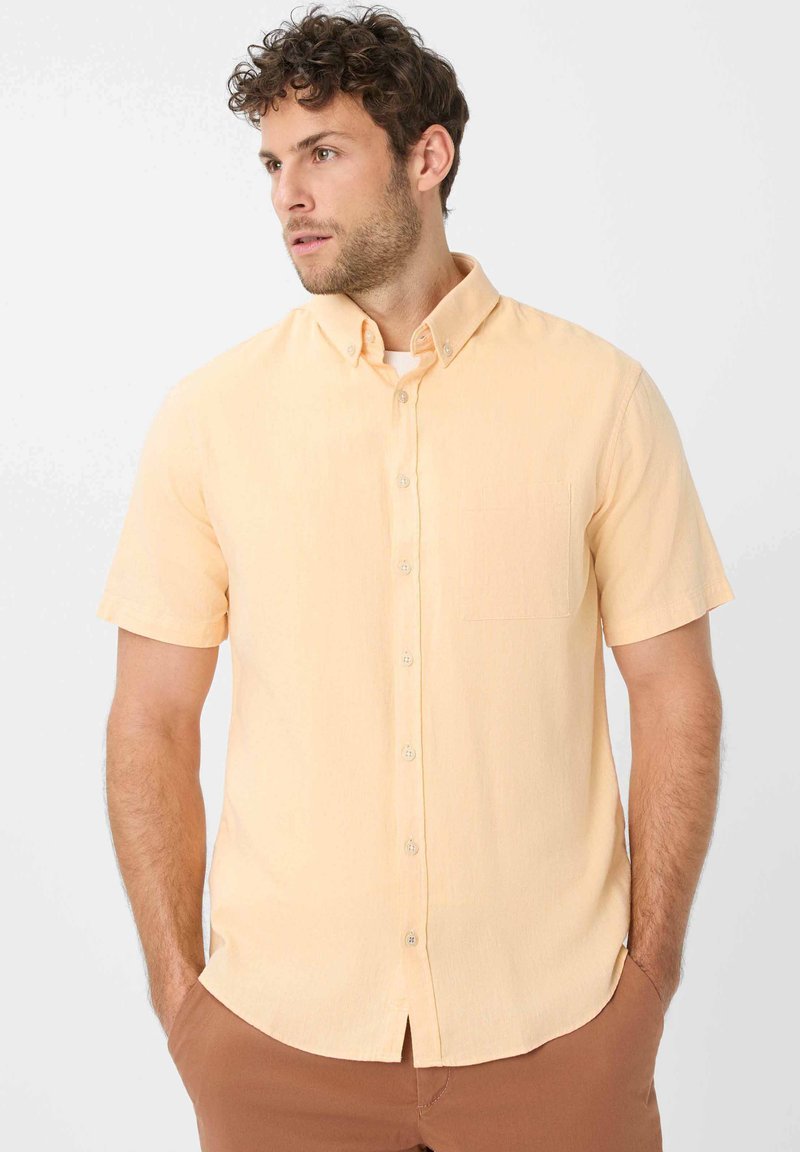 Korte mouwen, knoopsluiting shirt van zacht perzikkleurig materiaal, met een borstzak en een puntige kraag. De textuur lijkt glad en lichtgewicht.