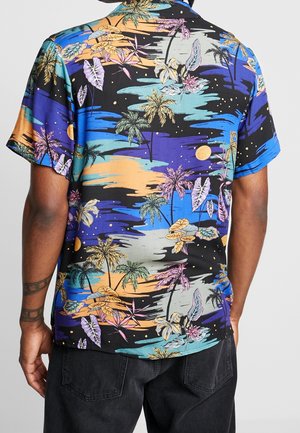 Vista trasera de una persona con una camisa de manga corta con un patrón de palmeras tropicales multicolores y flores sobre fondo negro.
