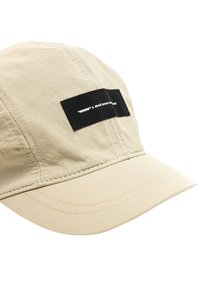 Gorra beige hecha de un tejido ligero, que presenta un parche rectangular negro con impresión blanca. La superficie tiene una textura enrejada y costuras visibles.
