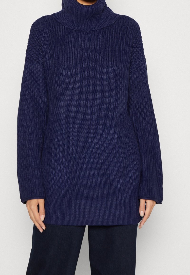 Pull à col roulé côtelé bleu marine à manches longues porté avec un pantalon foncé, les mains détendues le long du corps.