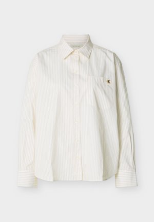 Fehér, hosszú ujjú, gombos ing vékony, függőleges csíkokkal, hegyes gallérral, mellső zsebbel és kis arany "cK" logóval a zsebén.