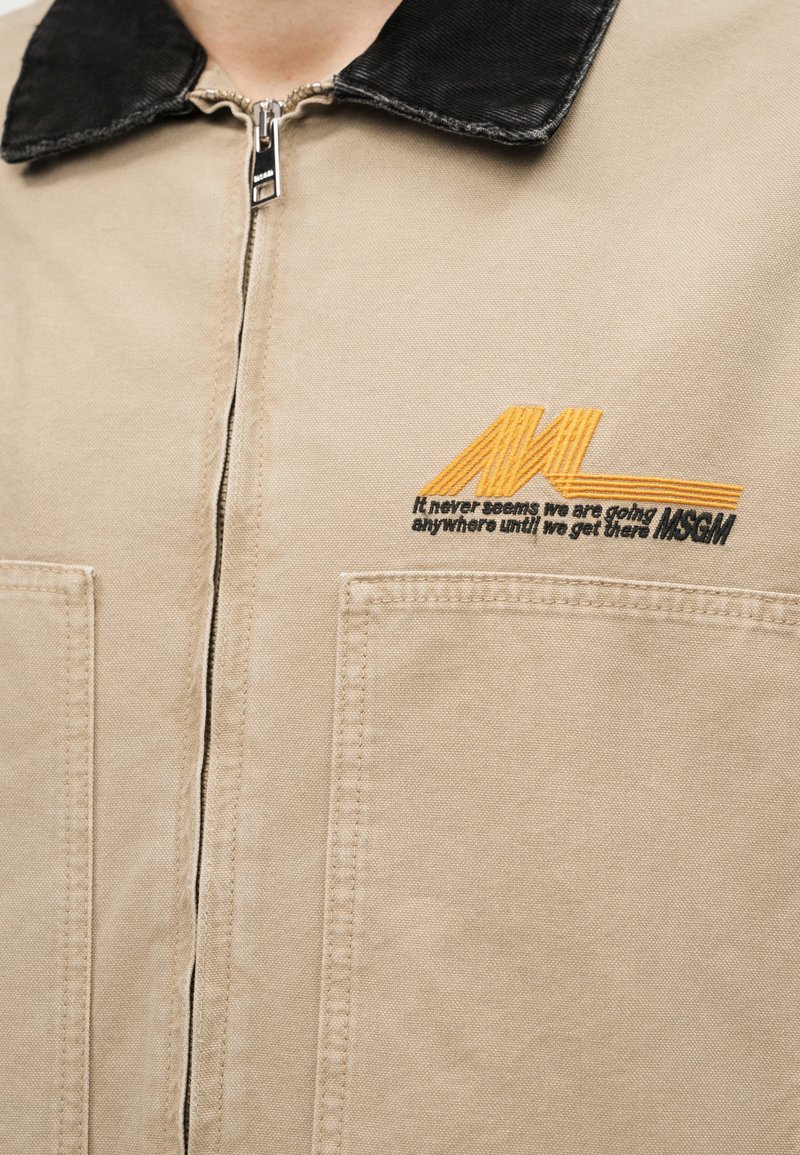 Beige jas met ritssluiting, zwarte kraag en voorzakken, met het oranje MSGM-logo en de tekst "Het lijkt nooit alsof we ergens heen gaan totdat we daar zijn."