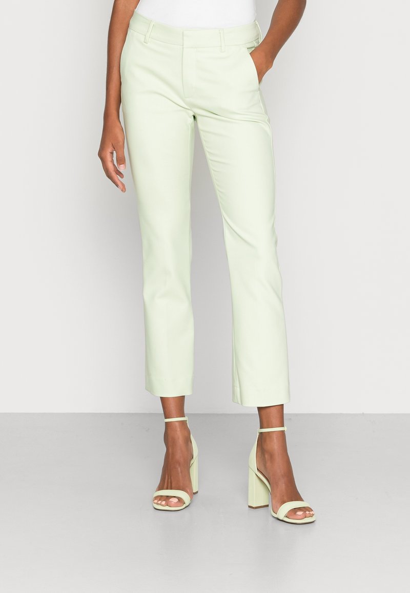 Femme portant un pantalon slim vert clair aux chevilles et des sandales à talons hauts ouvertes assorties, debout avec une main dans la poche.