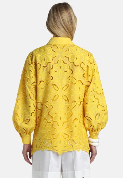 Sieviete, kas ģērbusies spilgti dzeltenā mežģīņu blūzē ar bumbieru piedurknēm un aproču aprocēm, valkā baltus šortus, redzama no aizmugures.