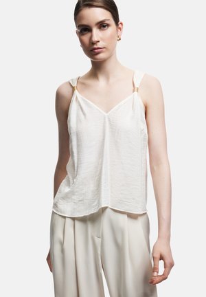 Jonge vrouw draagt een witte mouwloze getextureerde top met gouden ringdetails op de bandjes en losse crèmekleurige broek.