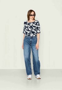 Marineblauwe bloes met korte mouwen en witte bloemen, gecombineerd met lichtblauwe rechte spijkerbroeken en witte sneakers met bruine zolen.