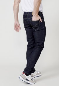 Jeans de mezclilla ajustados en azul oscuro con costuras amarillas, cinco bolsillos y una etiqueta de cuero marrón en la parte trasera de la cintura. Combinados con zapatillas en gris y blanco.