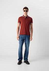 Burgundska polo majica s ovratnikom s bijelim prugama, uparena s plavim trapericama i crnim tenisicama. Model koji nosi sunčane naočale stoji uz jednostavnu pozadinu.