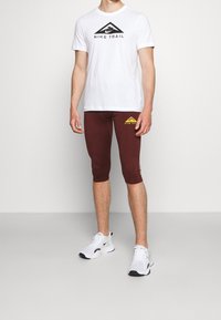 Camiseta Nike Trail blanca con logo negro, leggings ajustados burdeos con logo amarillo y zapatillas blancas con acentos negros. Tejido suave y ceñido al cuerpo.