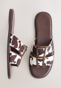 Pantoletten mit Kuhfellmuster, weichem, strukturiertem Obermaterial und brauner Gummisohle. Verfügen über zwei breite Riemen mit einem Kreuzdesign als Akzent.
