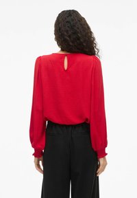 Blusa roja de manga larga con tejido texturizado, detalle de abertura en la parte posterior y puños fruncidos, combinada con pantalones anchos negros.