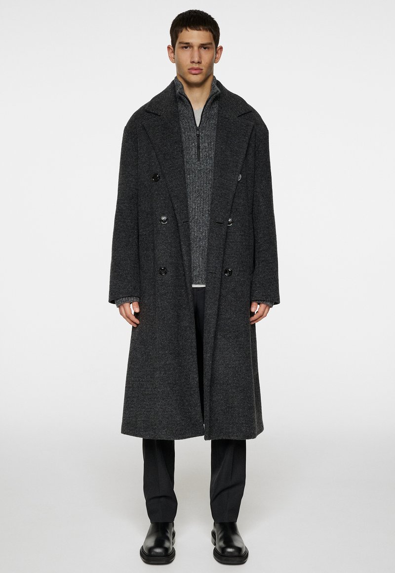Cappotto grigio in lana a doppio petto con ampi risvolti e bottoni neri, indossato sopra un maglione grigio con zip e pantaloni neri, completato da stivali neri.