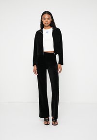 Sort velour cropped jakke med lynlås og matchende højtaljede bukser. Hvid tætsiddende crop top nedenunder. Model iført sorte hæle.
