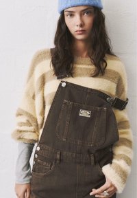 Braune Denim-Latzhose mit verstellbaren Trägern, Vordertasche und Metallbeschlägen, getragen über einem gestreiften beigefarbenen und cremefarbenen Pullover mit langen Ärmeln.