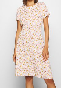 Robe florale rose à manches courtes, avec des motifs de fleurs blanches et jaunes sur un tissu texturé. Silhouette en A avec des détails de fronces subtils.