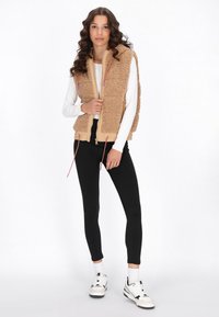 Beige, flauschige Weste mit einem Reißverschluss vorne und elastischem Saum, kombiniert mit einem langen weißen Hemd. Schwarze schmales Hosen und weiße Sneaker runden den Look ab.