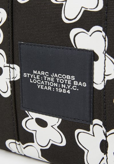 Sac fourre-tout en tissu noir avec des motifs floraux blancs. Une étiquette rectangulaire affiche "Marc Jacobs", "The Tote Bag", NYC, et "1984".