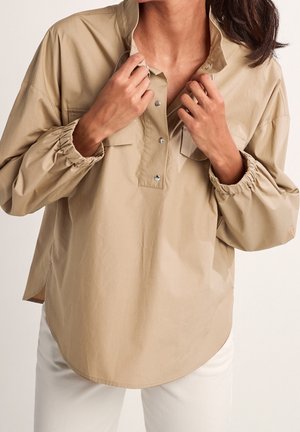 Femme ajustant le col d’une chemise beige ample à boutons avec des poignets élastiques, associée à un pantalon blanc sur un fond uni.