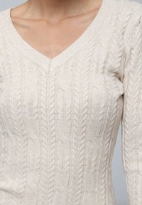 Pull en maille tressée de couleur beige clair. Présente un décolleté en V profond, des manches longues et un motif texturé avec des détails tressés.