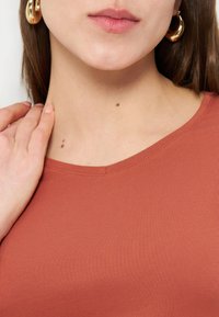 Γυναίκα που φορά χρυσούς κρίκους στα αυτιά και τοπ σε σκουροκέχρωμο πορτοκαλί, με δύο ελιές στο λαιμό, αγγίζοντας τον αριστερό ώμο με το αριστερό χέρι.