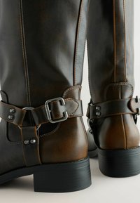 Kozaki skórzane sięgające do kolan w ciemnobrązowym odcieniu z jaśniejszymi akcentami, z regulowanymi metalowymi klamrami i teksturowanymi powierzchniami. Czarne obcasy.